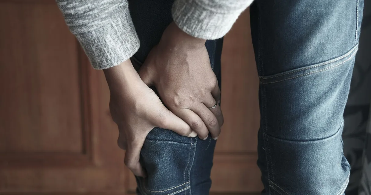 Homem de calça jeans segurando o joelho com as duas mãos por causa da dor